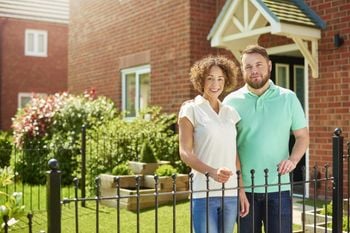 a mid adult couple stand at the front of their house and smile to camera . They are a mixed race couple , and their house is part of a new build housing development.