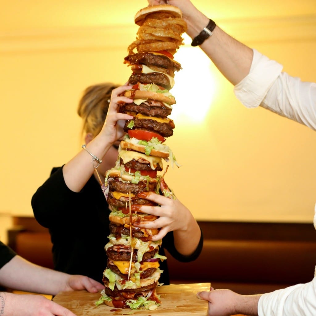 The UK's tallest burger is carefully carried to the bar at The George in Stockton. See SWNS story SWBURGER; A restaurant owner has created the daddy of all meals - Britain's tallest burger which is bigger than his DAUGHTER. The whopping two-and-a-half-foot '999 burger' is higher than Craig Harker's toddler and trumps the previous UK record by several inches. It gets its name from the 30,000 calorie content - enough to feed one person everyday for TWO WEEKS - which could leave any diner dialling for an ambulance. Regulars at The George in Stockton, Teeside, have been giving owner Mr Harker a big patty-on-the back for the 28 inch culinary masterpiece.