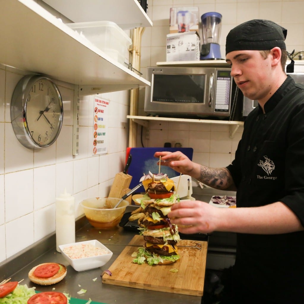 chef Josh Heslop, 19, starts the delicate process of building the UK's tallest burger. See SWNS story SWBURGER; A restaurant owner has created the daddy of all meals - Britain's tallest burger which is bigger than his DAUGHTER. The whopping two-and-a-half-foot '999 burger' is higher than Craig Harker's toddler and trumps the previous UK record by several inches. It gets its name from the 30,000 calorie content - enough to feed one person everyday for TWO WEEKS - which could leave any diner dialling for an ambulance. Regulars at The George in Stockton, Teeside, have been giving owner Mr Harker a big patty-on-the back for the 28 inch culinary masterpiece.