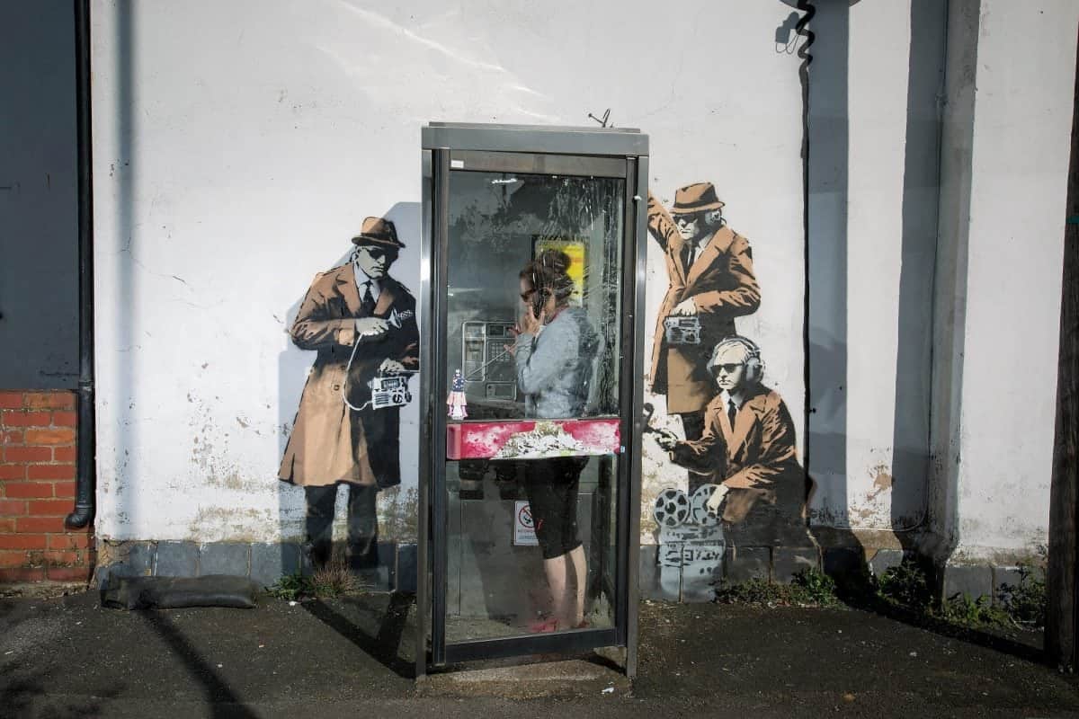 FILE PHOTOS OF phone box which has been surrounded by new Banksy artwork in Cheltenham. 14th April 2014. See SWNS story SWBANKSY; A famous Banksy once valued at a million pounds which "fell" off a wall appears to be for sale on Facebook. The elusive artist created 'spy booth' depicting three 'spooks' snooping on a phone box with listening devices near GCHQ in Cheltenham, Glos., in 2014. But it was reduced to a pile of rubble when builders worked on the wall above the artwork and it collapsed last summer. Building owner David Possee took the bits to the council for safekeeping, but yesterday (Weds) the broken pieces appeared for sale on Facebook.