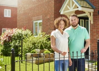 a mid adult couple stand at the front of their house and smile to camera . They are a mixed race couple , and their house is part of a new build housing development.